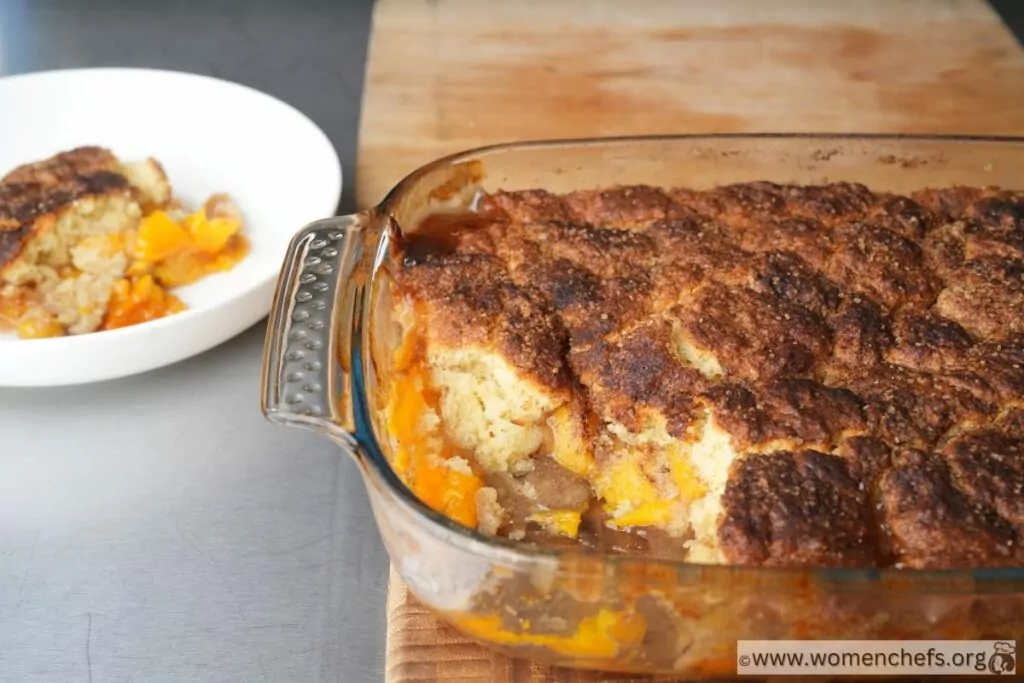 Cooked Peach Cobbler with a spoonfull taken from the tray and placed on a plate next to the tray